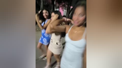 Media: A video captures three women dancing enthusiastically outdoors at night, wearing colorful, casual summer outfits. The background features blurred, dimly lit trees and a hint of pink.