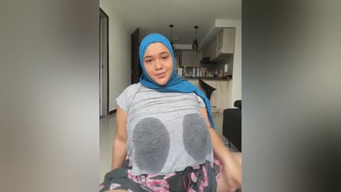 Media: Video of a smiling, light-skinned woman with a blue hijab and gray T-shirt featuring large, gray cartoon breasts. She wears floral pajama pants in a modern, open-plan kitchen.