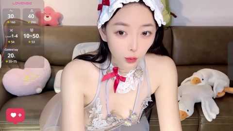 Media: Video of an Asian woman with long black hair, fair skin, wearing a revealing maid outfit with a lace choker, sitting on a brown sofa surrounded by stuffed animals.