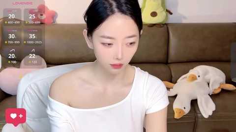 Media: Video of a young Asian woman with fair skin, dark hair tied back, wearing a white off-shoulder top, sitting on a brown couch. Background includes plush toys, a white and brown stuffed dog, and a green stuffed toy.