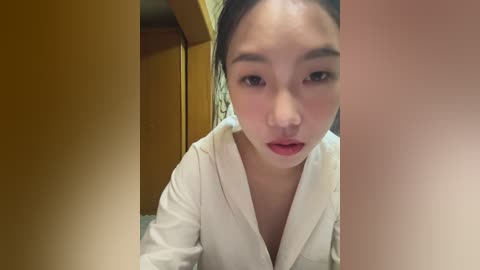 Media: Video of a young Asian woman with fair skin, dark hair in a bun, wearing a white bathrobe, looking contemplative. Background features wooden walls and a blurred wooden door.