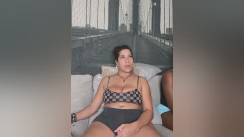 Media: Video of a plus-sized woman with dark hair, wearing a checkered bra and high-waisted shorts, sitting on a beige couch in front of a large black-and-white mural of the Brooklyn Bridge.