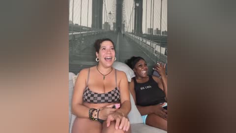 Media: Video of a cheerful woman with medium build, light skin, and dark hair, wearing a black-and-white checkered bikini, sitting next to a dark-skinned woman with a medium build, wearing a black tank top and shorts, both smiling.