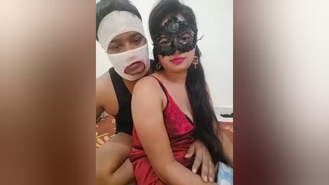 Media: Video of two women: one with a white bandage over her face, wearing a black mask, red dress, and gold earrings, the other with a black mask, red lipstick, and long black hair, seated on a bed.