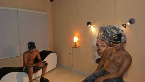 Media: Video of a small, dimly-lit room with a shirtless man wearing camouflage shorts sitting on a white couch, a man with a headband and tattoos sitting next to a large, colorful portrait of a woman on a beige wall.