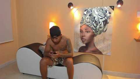 Media: Video of a shirtless Black man in patterned shorts, sitting on a white sofa with a large, colorful African-inspired portrait on the wall.