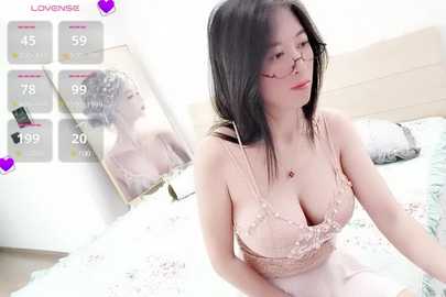 Media: Video of an Asian woman with long black hair, wearing glasses and a lacy pink bra, sitting on a bed in a bright, modern bedroom.