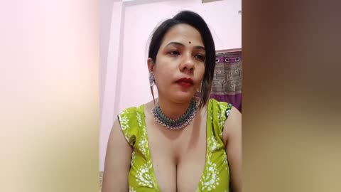 Media: Video of a South Asian woman with medium skin tone, black hair, and a bindi, wearing a green floral top with a deep V-neck, a silver necklace, and a subtle makeup look.