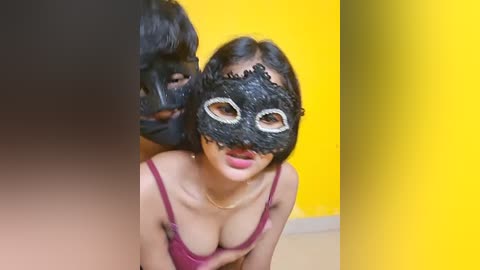 Media: Video of a young Asian woman with fair skin, wearing a black Venetian mask with intricate white designs, and a red top. A man behind her has a similar mask on. Background is a yellow wall.