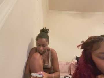 Media: Video of a young woman with dark skin and curly hair tied in a bun, wearing a beige tank top, sitting on a bed against a beige wall, looking down at a pink object.