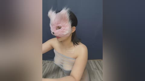 Media: Video of an Asian woman with medium skin tone, wearing a sheer, light grey bodysuit and a pink feathered mask, sitting on a wooden floor against a dark blue backdrop.