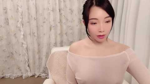 Media: Video of an East Asian woman with fair skin, dark hair in a bun, wearing a sheer, off-shoulder white top. She sits on a white chair with a floral patterned cushion. The background features white lace curtains and a beige floor.