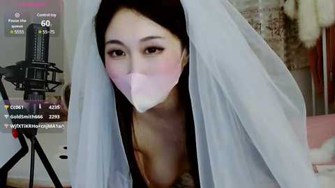 Media: Video of a woman with a mask, wearing a white veil, in a room with a green screen, a camera on a tripod, and a cityscape poster in the background.