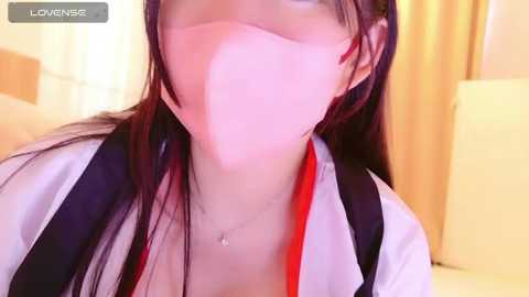 Media: A close-up video of an Asian woman with long black hair, wearing a white mask, red top, and black suspenders, indoors against a yellow curtain background.