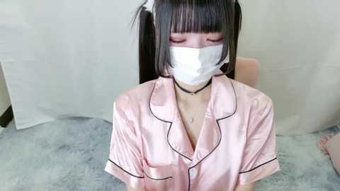 Media: A video of an Asian woman with a fair complexion, wearing a pink satin robe, a white mask, and black choker, kneeling on a light grey carpet.