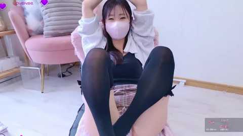 Media: Video of an East Asian woman with long black hair, wearing a schoolgirl outfit, mask, and thigh-high stockings, sitting on the floor with legs raised, in a pink room.