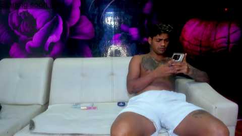 Media: Video of a shirtless, tattooed man with medium skin tone, wearing white shorts, sitting on a white couch, engrossed in a smartphone, in a dimly lit room with purple floral wall art.