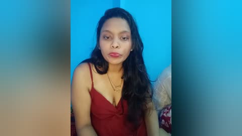 Media: Video of a young South Asian woman with long, wavy black hair, wearing a red camisole, standing against a blue wall, holding a heart-shaped necklace, in a dimly lit room.