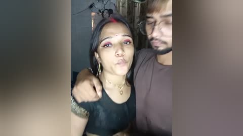 Media: Video of a South Asian woman with fair skin, red bindi, and red eyeshadow, wearing a black top and gold bangles, holding a man's hand. Background shows a dark room with indistinct objects.