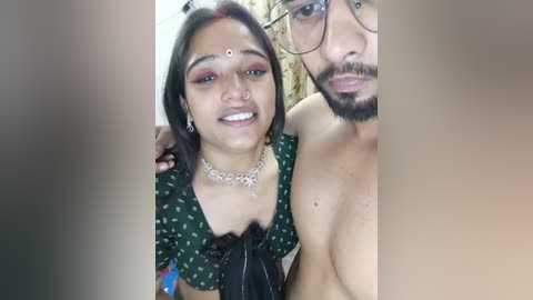 Media: A video shows a smiling South Asian woman with dark hair, wearing a green blouse with a black bow and silver jewelry, and a shirtless South Asian man with a beard.