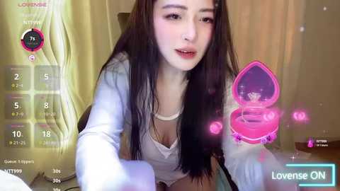 Media: Video of a young East Asian woman with long, straight black hair, wearing a white top, holding a pink heart-shaped box. Background shows a cozy room with yellow curtains.