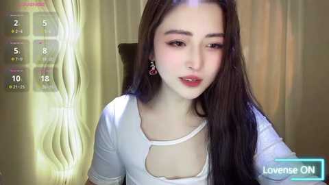 Media: A video of an Asian woman with long brown hair, fair skin, and red lipstick, wearing a white top with a cutout design, sitting indoors with a cream-colored curtain and a blue and white digital display.