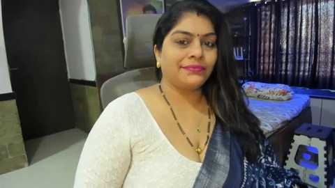 Media: Video of a South Asian woman with medium skin tone, long black hair, wearing a white lace blouse, dark blue saree, gold necklace, sitting on a bed with patterned blanket, in a room with wooden furniture and window curtains.