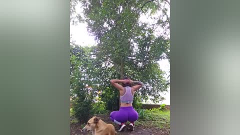 Media: Video of a woman squatting, stretching, wearing a purple sports bra and leggings, in a lush, green forest setting with a beige dog resting beside her.