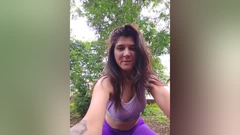 Media: Video of a young woman with long brown hair, wearing a purple sports bra and purple leggings, posing in a forest with lush green foliage and a tree in the background.