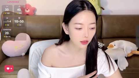 Media: Video of a young East Asian woman with long black hair, fair skin, and light makeup, wearing a white off-shoulder top, sitting on a brown couch with plush toys and a green bag in the background.