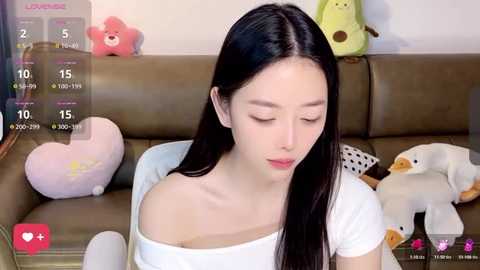 Media: Video of an Asian woman with long black hair, wearing a white top, sitting on a brown leather sofa with stuffed animals, including a pink bear, in a cozy room.