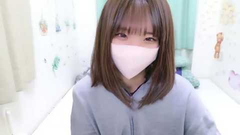 Media: Video of an Asian woman with shoulder-length brown hair, wearing a light blue scrub top and a white surgical mask, standing in a sterile, white-tiled hospital room with pastel-colored decor and medical equipment in the background.