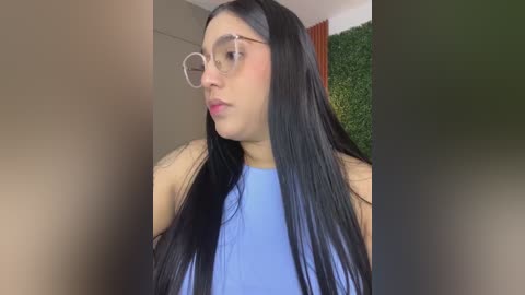 Media: Video of a young woman with long black hair, wearing glasses and a light blue sleeveless top, standing against a green wall with a brown door and orange curtain in the background.