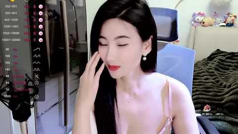 Media: A video of a young East Asian woman with long black hair, fair skin, and red lipstick, wearing a pink lace bra, in a cozy bedroom with a blue chair and stuffed toys.