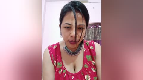 Media: Video of an Indian woman with medium skin tone, black hair, and a red blouse adorned with green leaves, wearing a silver necklace. Background features a pink wall and a partially visible curtain.