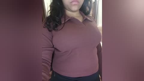 Media: Video of a woman with medium skin tone and wavy dark hair, wearing a tight, maroon polo shirt and black pants, standing in front of a maroon wall.