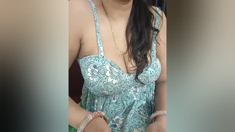 Media: Video of a South Asian woman with medium skin tone and long black hair, wearing a light blue, floral-patterned, sleeveless top with spaghetti straps, holding a green bag.