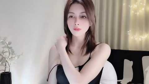 Media: Video of an East Asian woman with straight, shoulder-length brown hair, fair skin, and light makeup, wearing a black spaghetti strap top, sitting indoors with a minimalist background featuring white curtains and a lit candle.