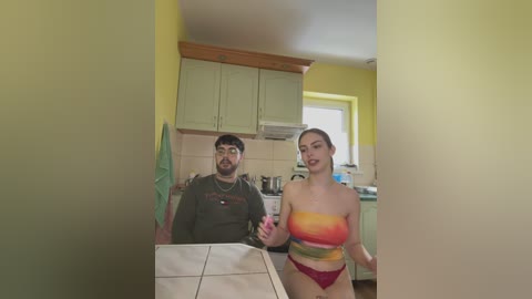 Media: Video of a man with a beard and glasses, and a woman with long brown hair, both in casual clothes, standing in a kitchen with yellow walls, cabinets, and a window.