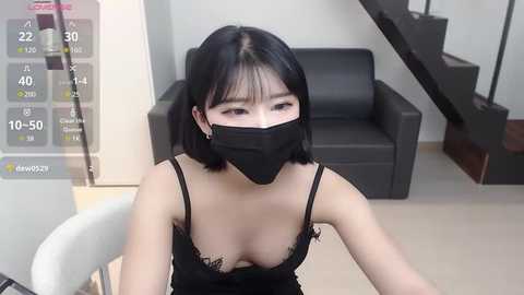 Media: A video of an East Asian woman with straight black hair and a black face mask, wearing a black lace bra, sitting on a grey chair in a modern living room.