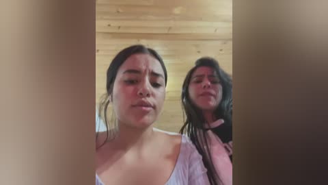 Media: Video of two young women with long dark hair, one wearing a white shirt, the other a pink sweater, standing indoors against a wooden wall.