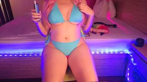 Media: Video of a young woman with long pink hair, wearing a light blue bikini, sitting on a bed with neon lights and stuffed toys, holding a phone.