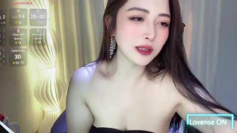 Media: Video of an East Asian woman with long, straight black hair, fair skin, and wearing a strapless black dress with silver earrings. She appears relaxed, with a soft smile. Background shows a modern, minimalist room with a digital clock and a greenish-yellow wall.