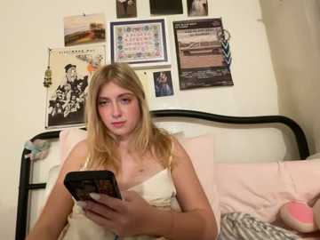 Media: Video of a young woman with blonde hair, fair skin, and a slender physique, wearing a white tank top, sitting on a bed with pink bedding, reading a smartphone, surrounded by framed photos and posters on a white wall.