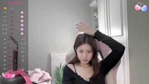 Media: Video of an Asian woman with long brown hair, wearing a black off-shoulder top, sitting in a pink gaming chair, scratching her head, surrounded by a cluttered desk with a pink towel, green plant, and a black cup.