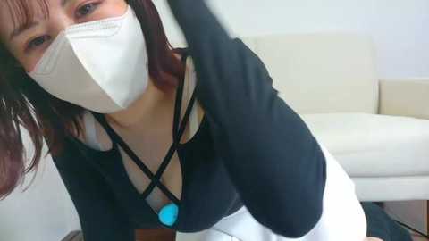 Media: A video of a woman in a black shirt, white pants, and a blue heart pendant necklace, wearing a white mask, taken from a low angle in a minimalist living room with white furniture.