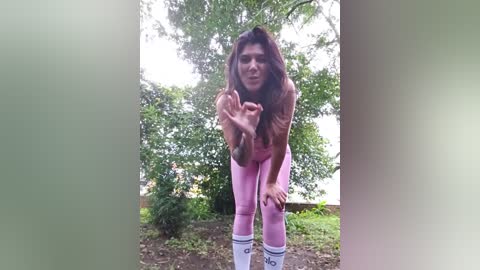 Media: Video of a woman with long brown hair, bending over, wearing pink leggings and white socks, in a green, leafy outdoor setting.