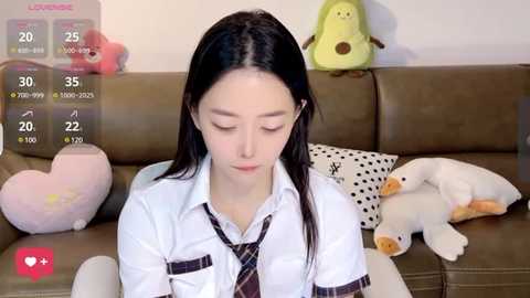 Media: Video of an Asian woman with long black hair, wearing a white blouse and a plaid tie, sitting on a brown couch with plush toys and a green frog plushie.