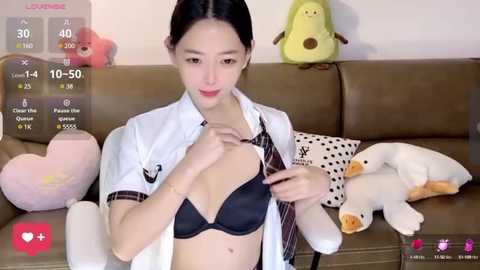 Media: Video of an East Asian woman with fair skin, long black hair, wearing a white shirt and black bra, adjusting her tie. Background includes a brown couch with plush toys, including a green frog.