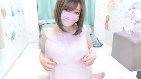 Media: A video of an East Asian woman with straight, shoulder-length brown hair, wearing a light pink dress and face mask, kneeling in a room with white walls and floral patterns.